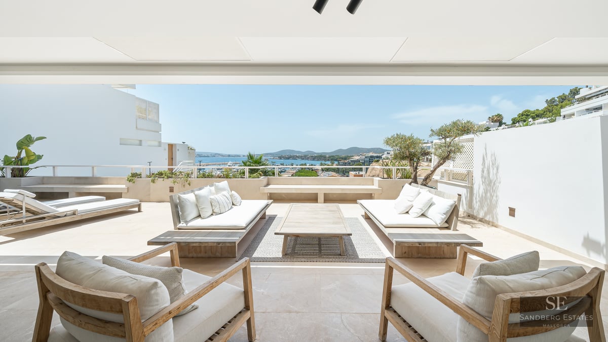 Spacious white terrace with wooden sofas and armchairs overlooking a blue sea and coastline under a bright sky.