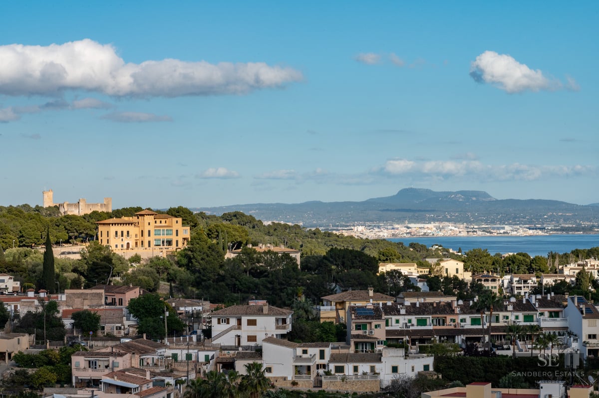 Panoramic landscape view showing a yellow villa, a historical stone castle on a hill, and the Mediterranean Sea in the distance.