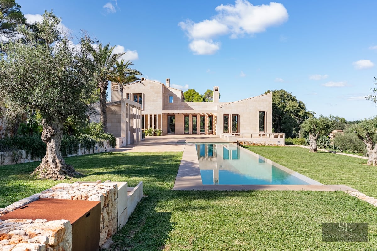 Modern sandstone villa with a long rectangular lap pool, lush green lawn, and ancient olive trees under a blue sky.