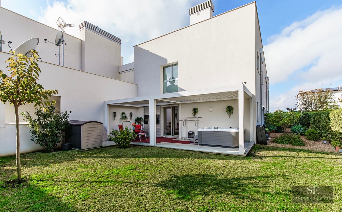 A modern white building with a green lawn and a covered terrace area featuring a hot tub and dining set.