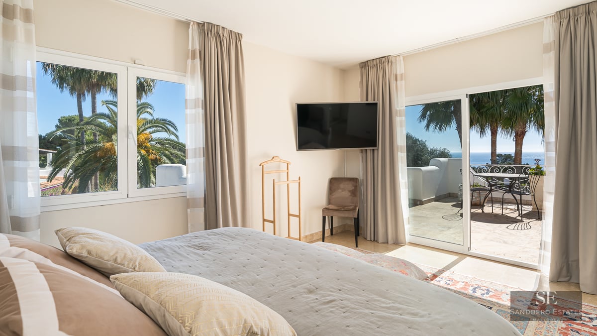 Modern master bedroom with large bed, wall-mounted TV, and sliding glass doors opening to a sunlit terrace with sea views.