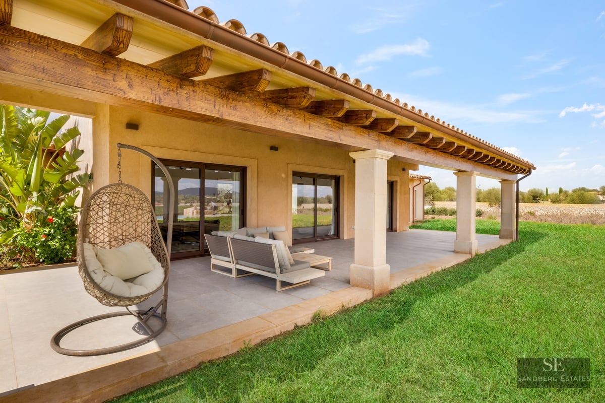 A covered terrace with rustic wooden beams, modern outdoor furniture, a hanging chair, and a green lawn under a blue sky.