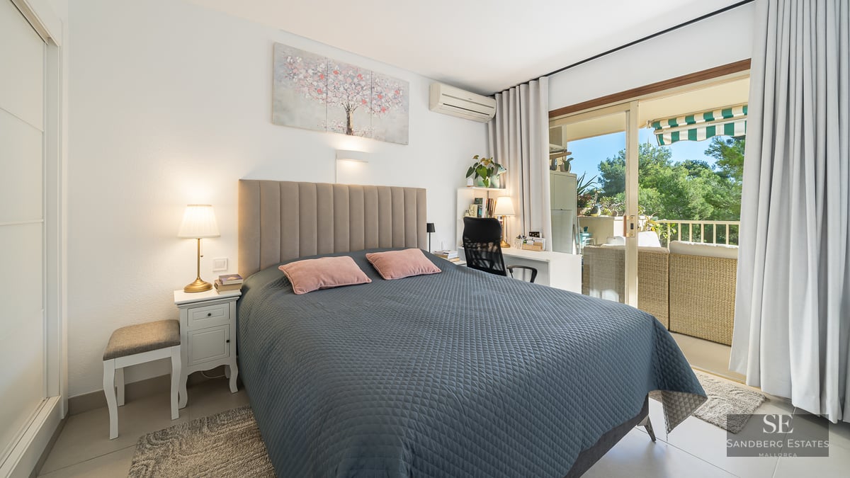 Chambre lumineuse avec lit matelassé gris, bureau et baies vitrées coulissantes ouvrant sur un balcon ensoleillé.