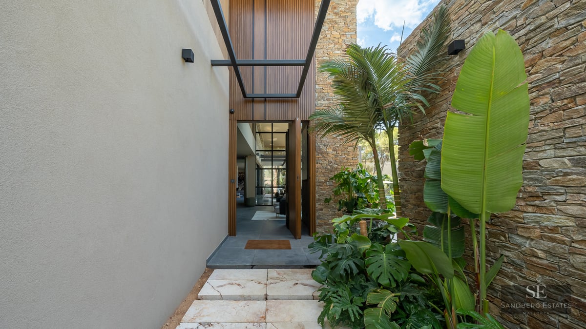 Stone path leading to a modern villa entrance with tropical plants, stone walls, and wooden door.