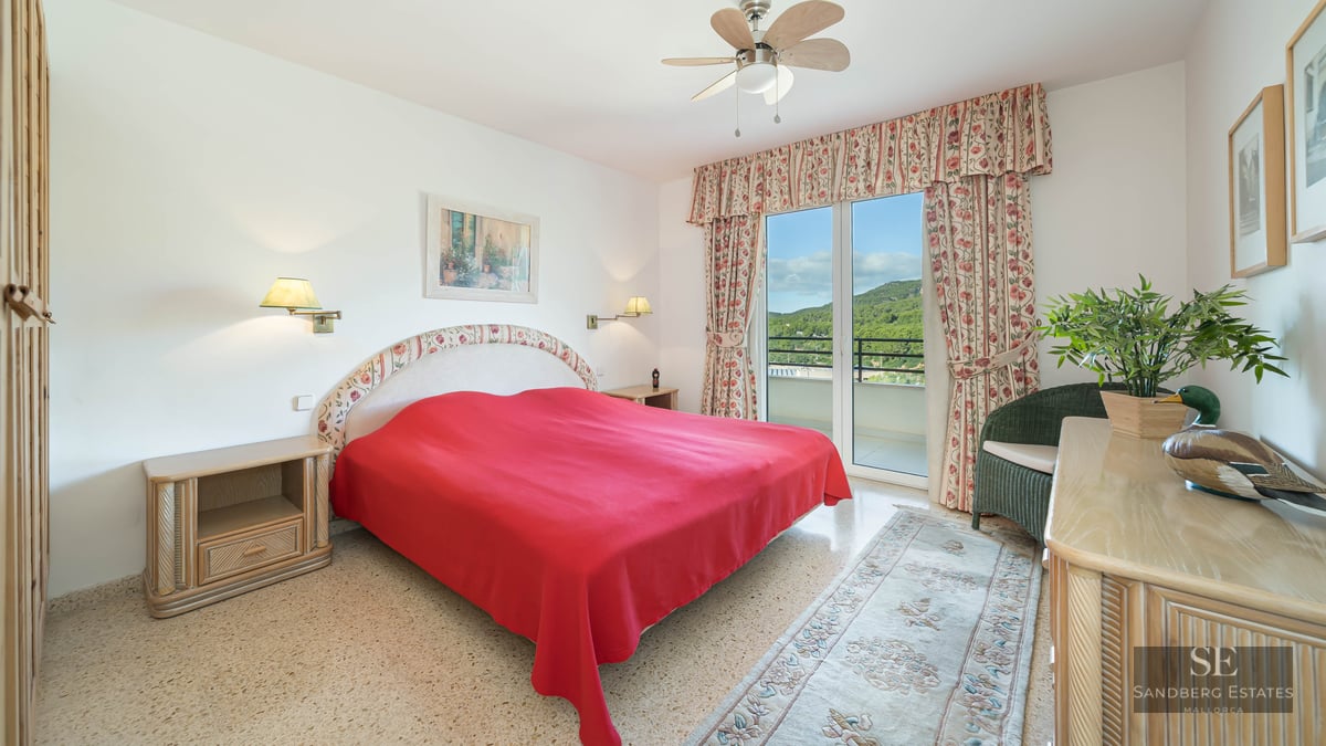 Bright bedroom featuring a red bedspread, floral curtains, and glass doors opening to a balcony with green hill views.
