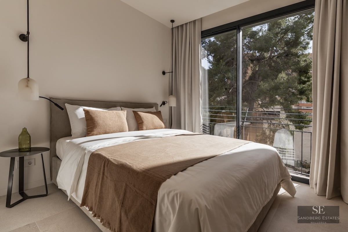 Bright bedroom with a large bed, neutral linens, designer pendant lights, and a floor-to-ceiling window viewing trees.