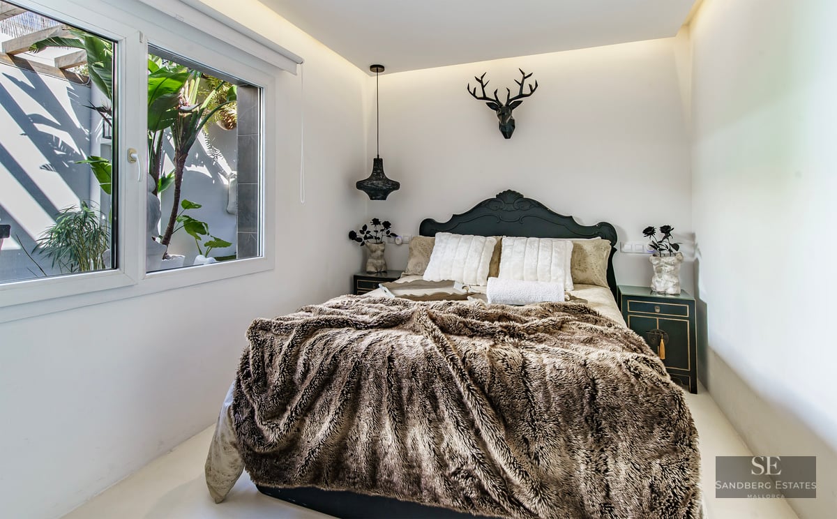 Bedroom with a brown faux fur throw, dark headboard, deer head wall sculpture, and window looking onto tropical plants.