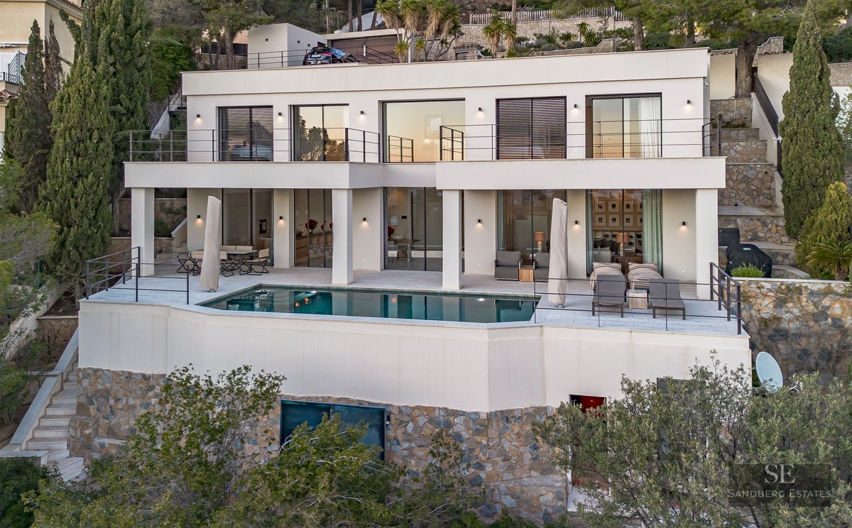 Two-story modern white villa with an infinity pool on a stone-clad terrace overlooking a hillside.