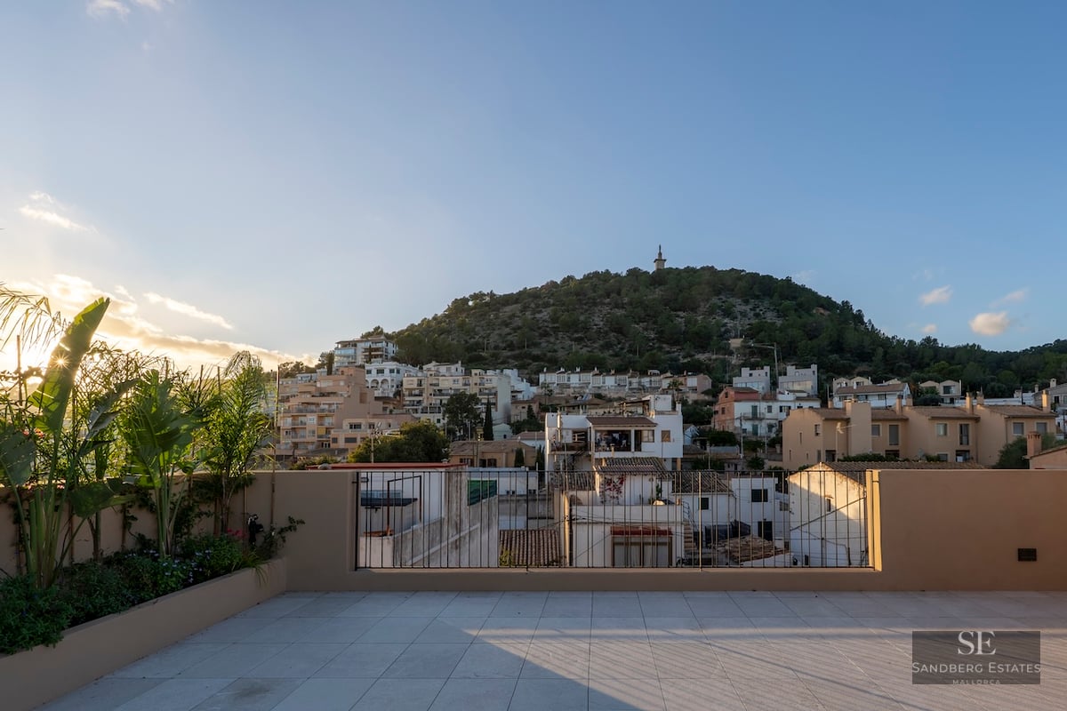 Grande terrasse carrelée avec garde-corps métallique offrant une vue sur un village méditerranéen et une colline au coucher du soleil.