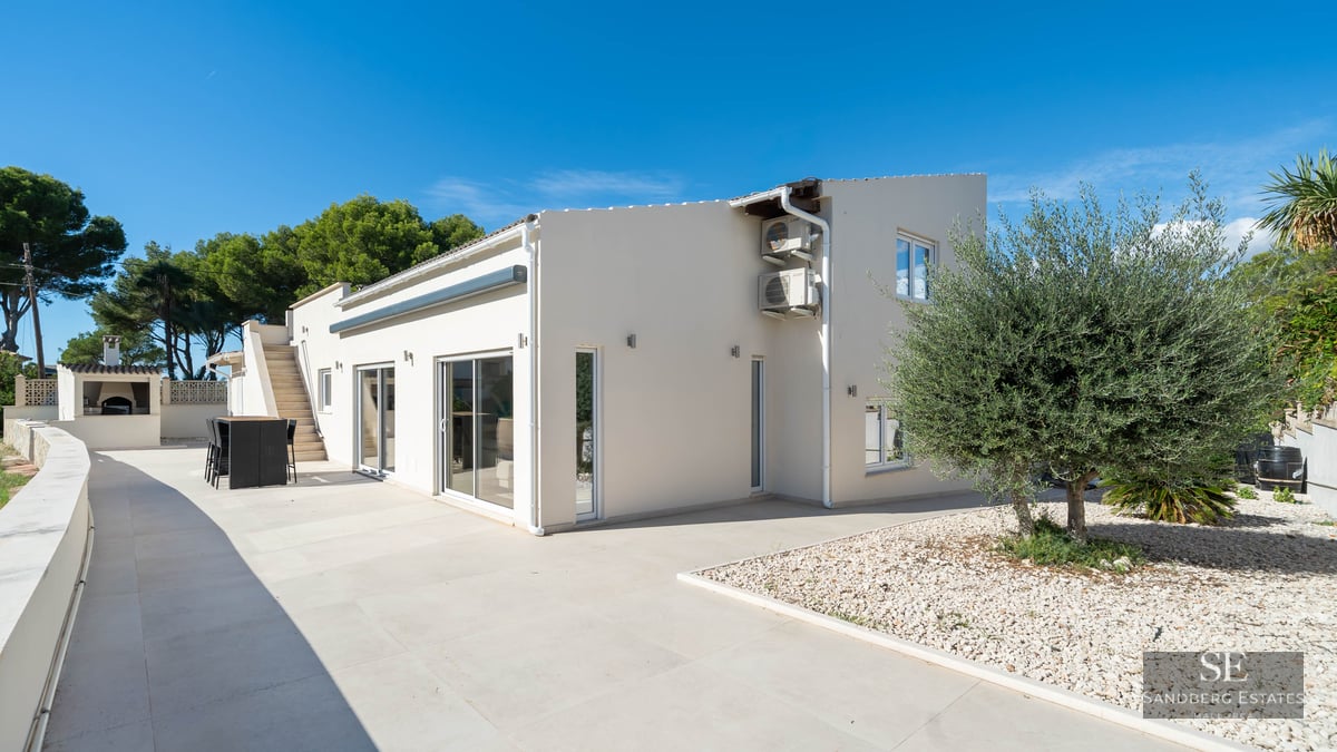 Grande terrasse en pierre blanche avec table à manger, olivier et façade de villa moderne sous un ciel bleu dégagé.