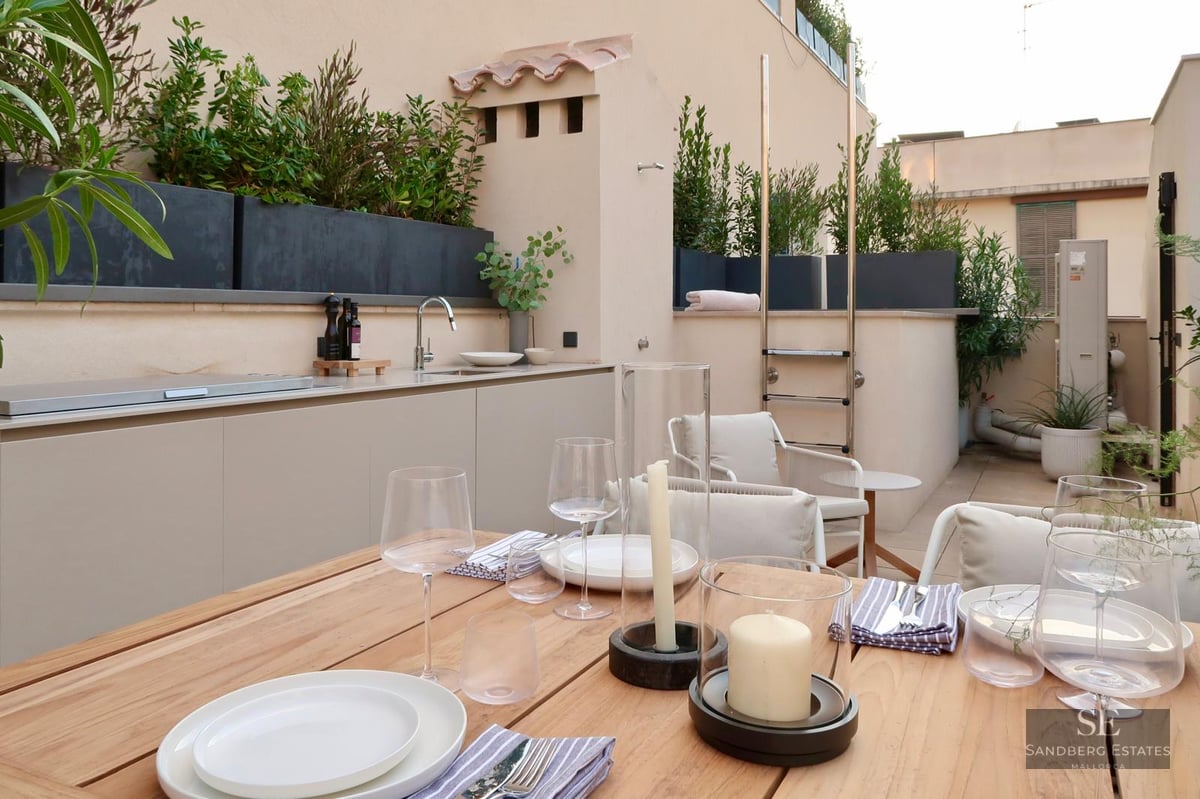 Wooden dining table on a modern terrace featuring an outdoor kitchen and lush greenery.