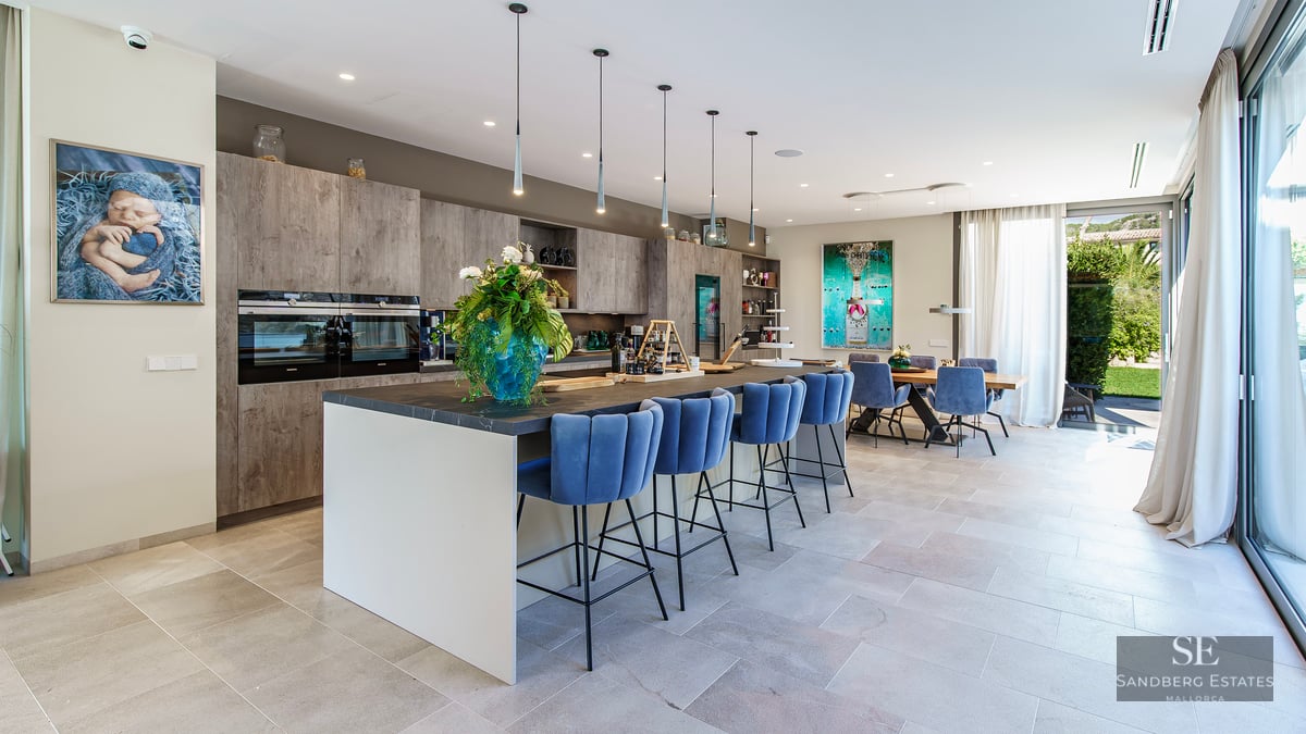 Cuisine moderne avec îlot central, tabourets en velours bleu et salle à manger intégrée baignée de lumière.
