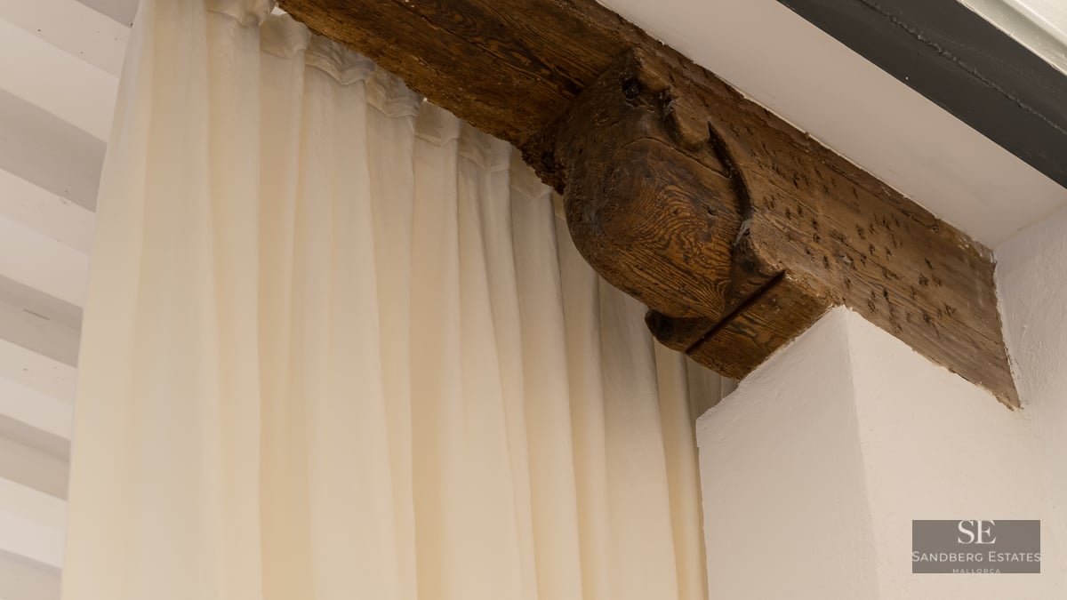 Close-up of a rustic carved wooden beam next to white curtains and a plaster wall.