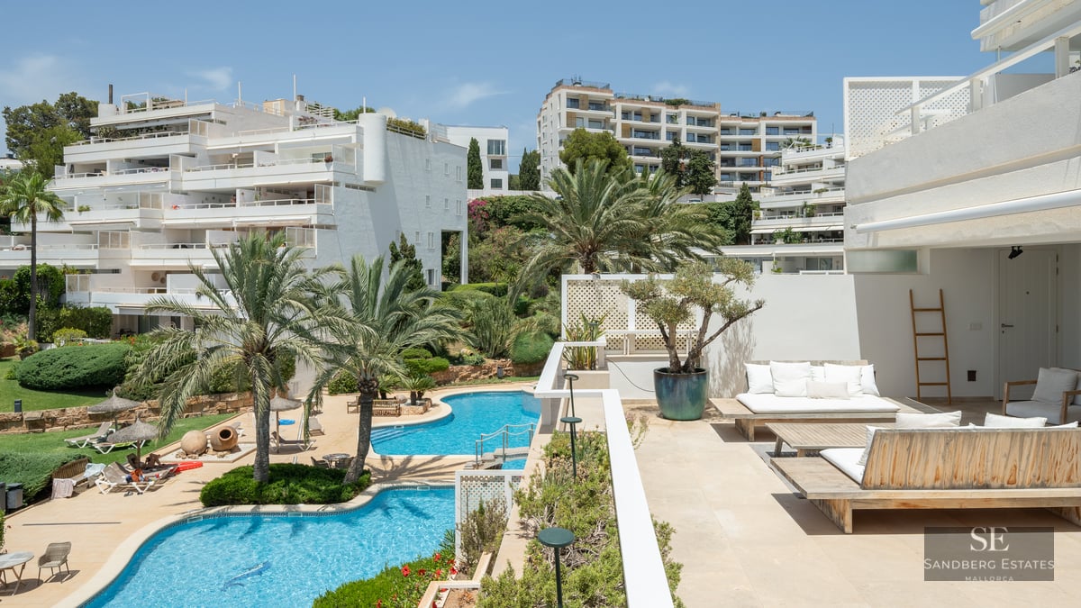 Sunny terrace with wooden lounge furniture overlooking multiple blue swimming pools and white Mediterranean buildings.