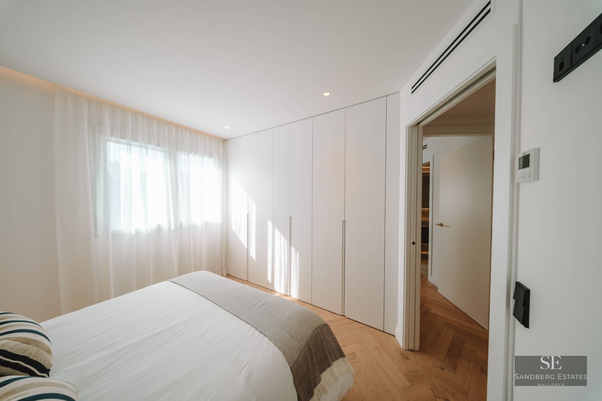 Bright bedroom featuring herringbone wood floors, floor-to-ceiling white wardrobes, a bed with white linens, and sheer curtains.