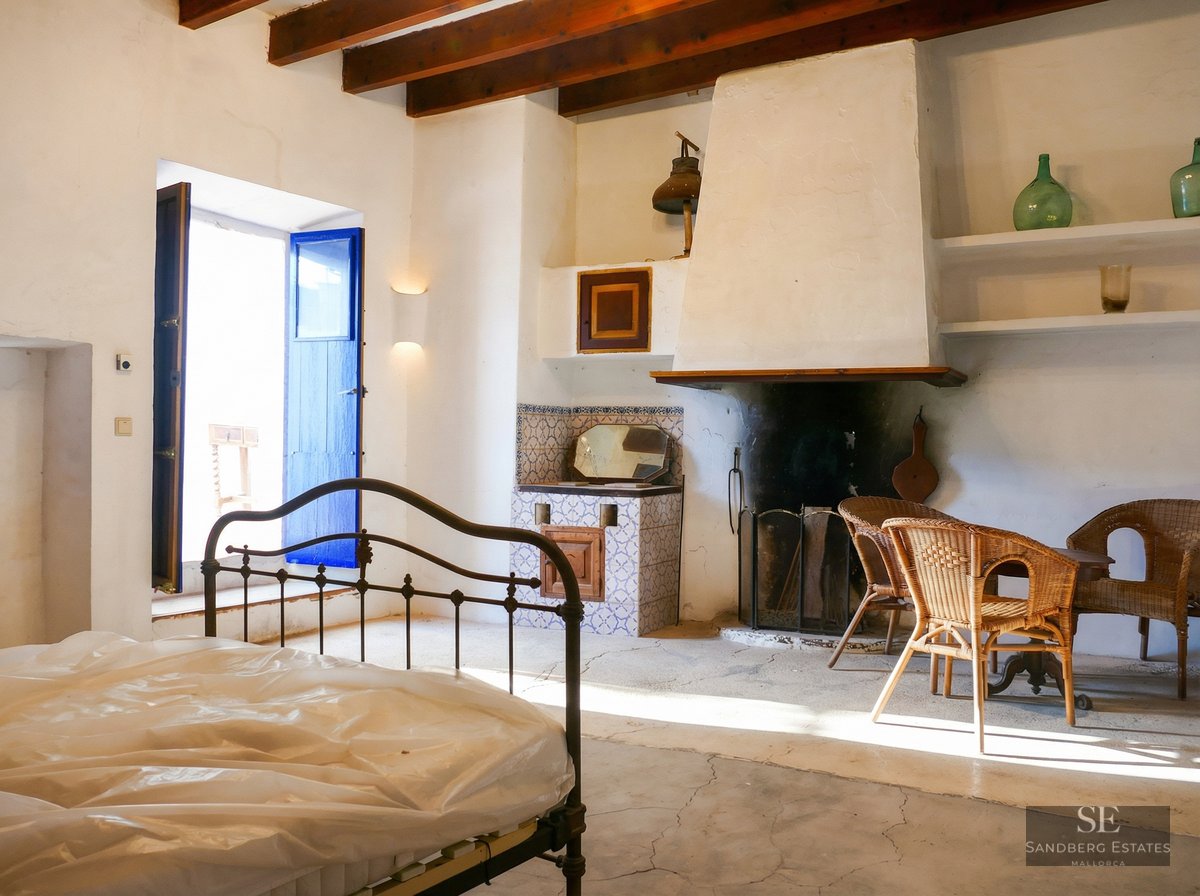 Bedroom with a wrought iron bed frame, a large white fireplace, blue wooden shutters, and wicker chairs under wooden beams.