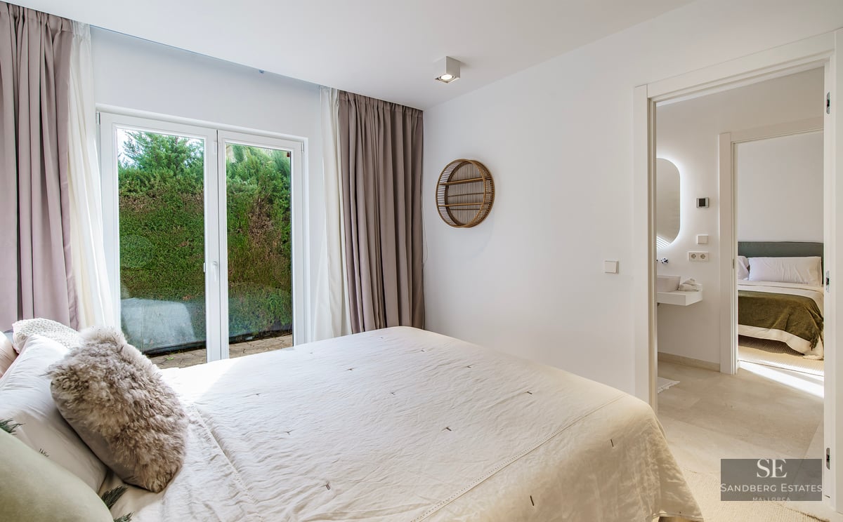 Bright master bedroom with white linens, glass doors to a garden, and a view into an ensuite bathroom.