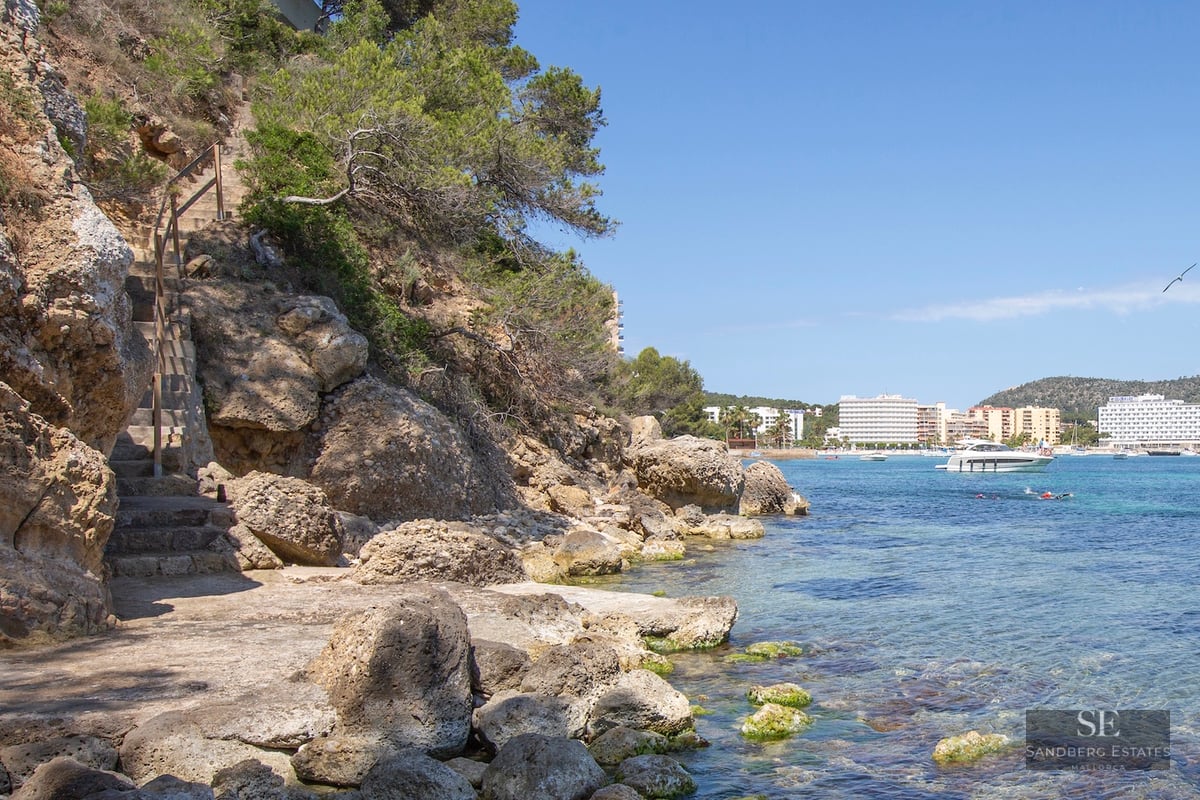 Stone stairs leading down a rocky cliff to turquoise sea with boats and buildings in the distance.