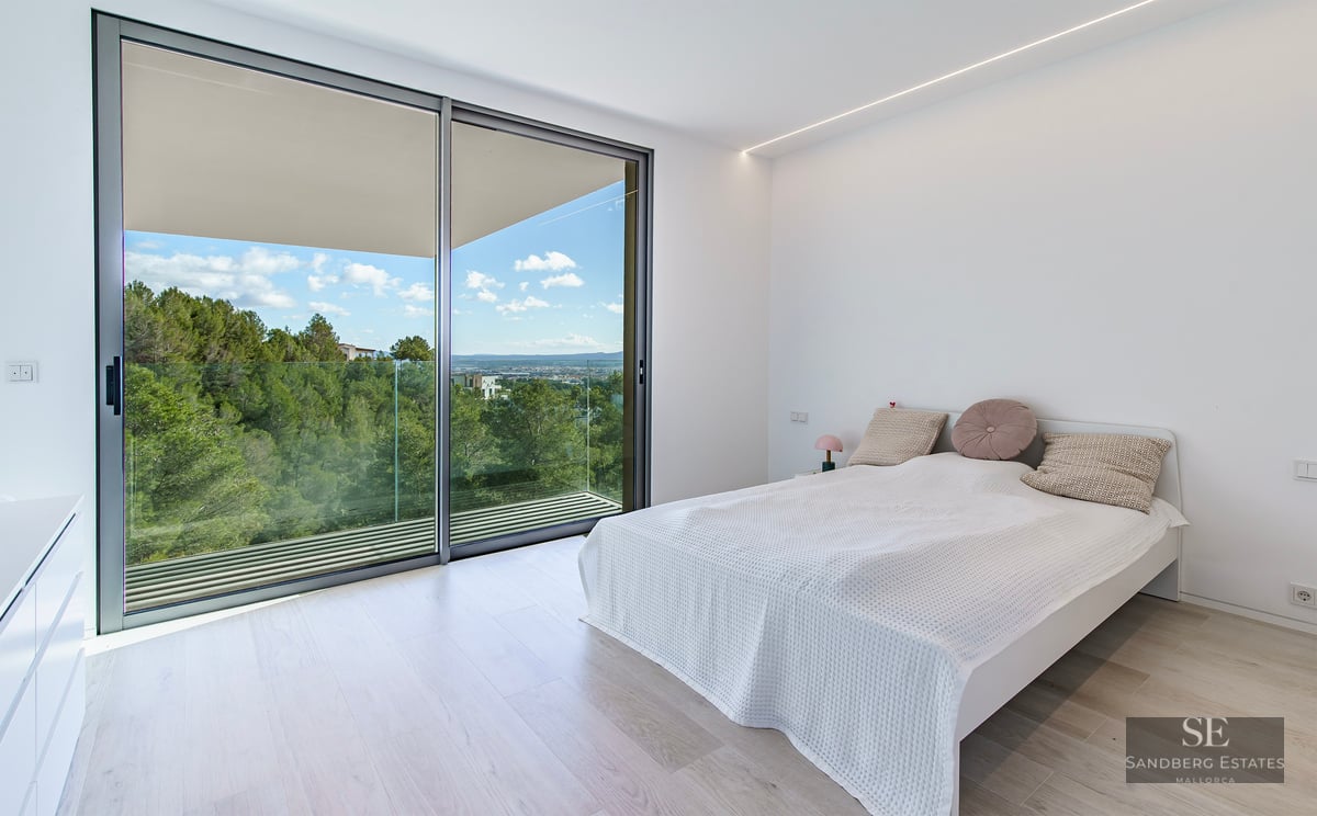 Bright minimalist bedroom with large glass sliding doors opening to a balcony with views of green hills and a blue sky.