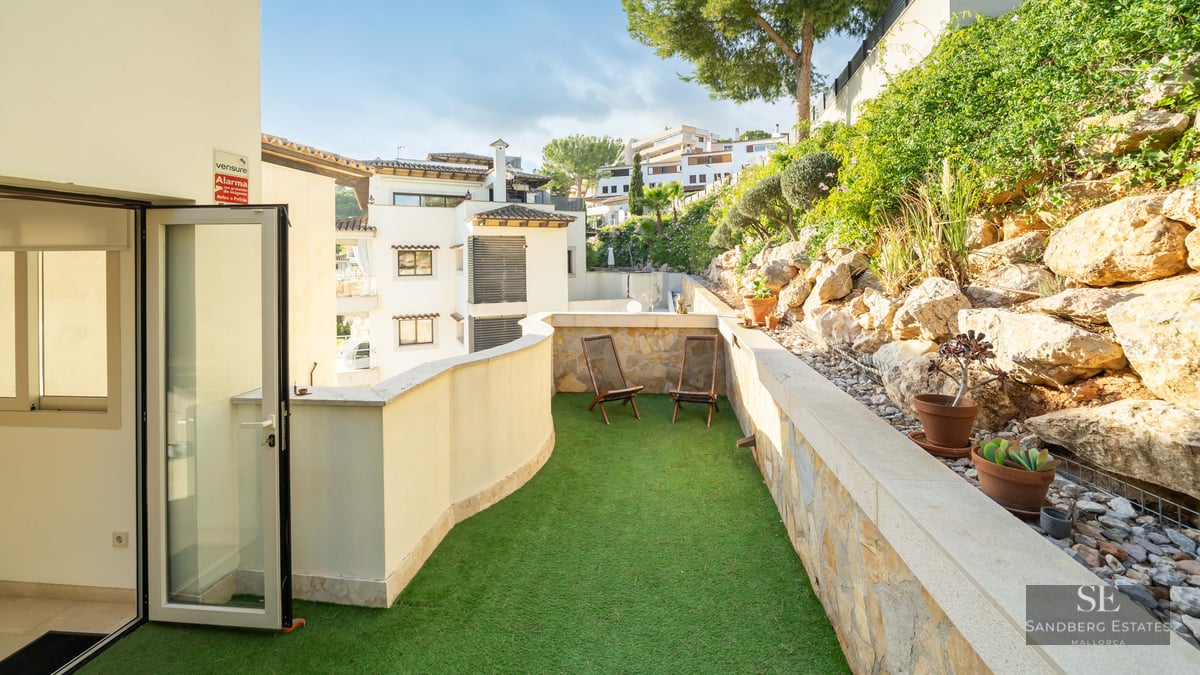 Sunlit terrace with green artificial grass, two wooden lounge chairs, and a natural stone wall.