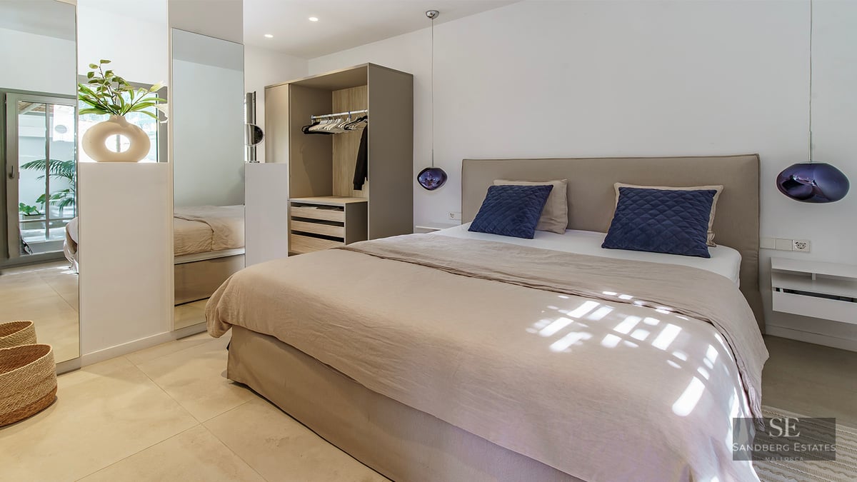 Bright modern bedroom featuring a beige bed, navy pillows, purple glass pendant lamps, and a built-in wardrobe with mirrors.