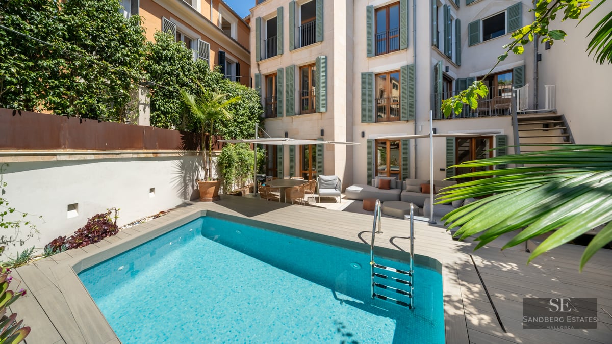 Light blue pool in a courtyard with wooden decking, outdoor sofas, and a building facade with green shutters.