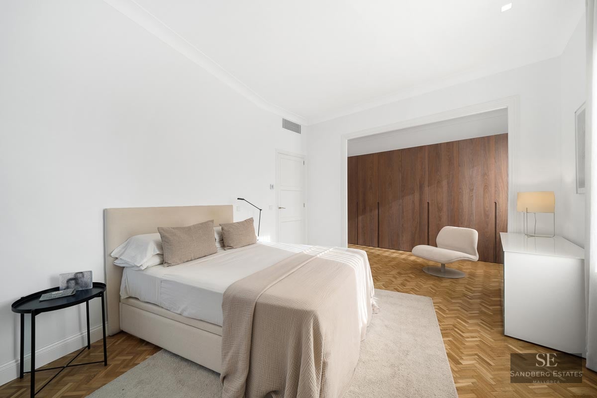 Bright minimalist master bedroom featuring a large bed, herringbone wood floors, and floor-to-ceiling wooden wardrobes.