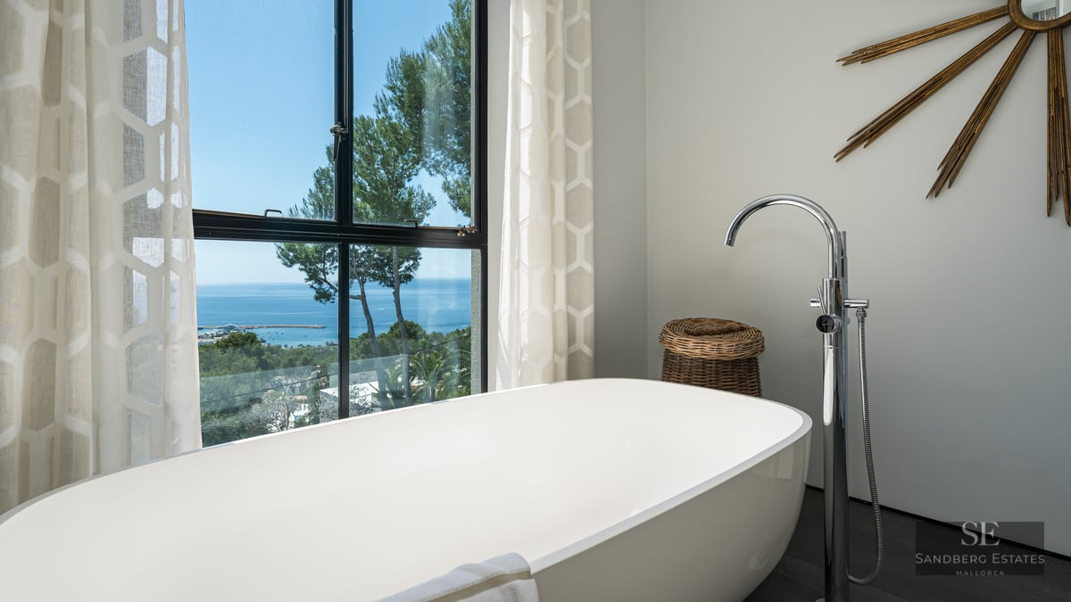 A white freestanding bathtub positioned in front of a large window overlooking the Mediterranean Sea and trees.