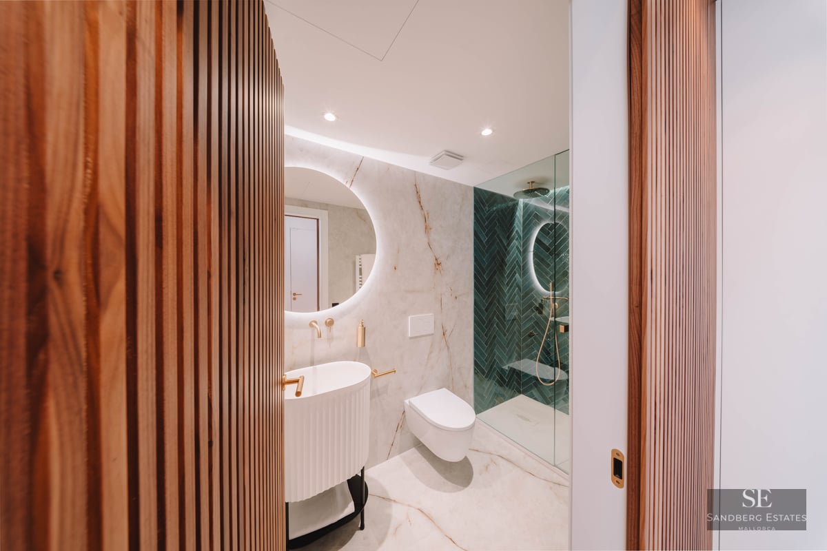 Modern bathroom featuring marble walls, a backlit circular mirror, gold fixtures, and green herringbone shower tiles.