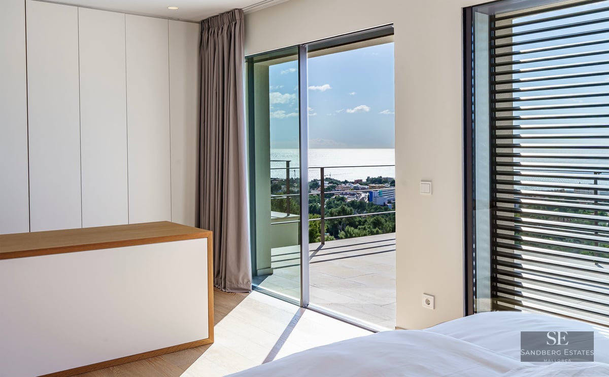Bright modern bedroom with floor-to-ceiling sliding glass doors opening to a terrace overlooking the ocean.
