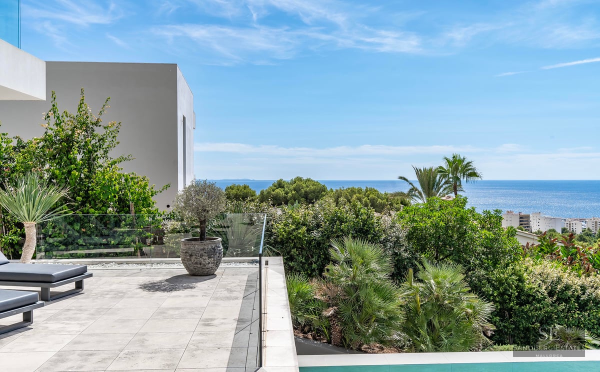 Modern stone terrace with glass railing, sun loungers, and lush green plants overlooking the blue Mediterranean Sea.