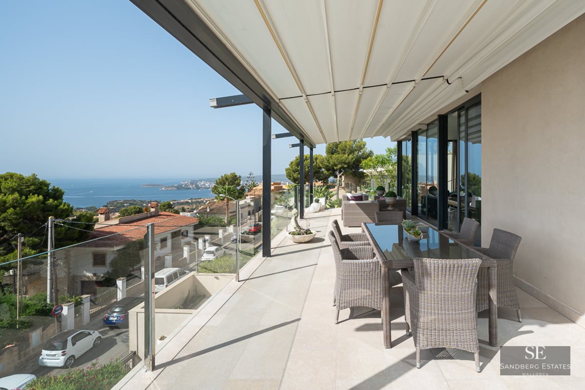 Spacious modern terrace with wicker dining furniture, glass railings, and expansive views of the ocean and coastal town.