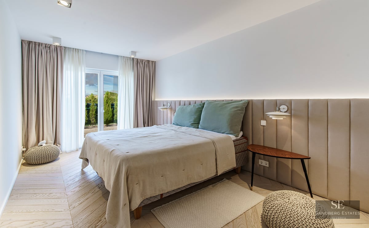 A bright modern bedroom featuring a large bed with a beige upholstered wall headboard and light wood chevron floors.