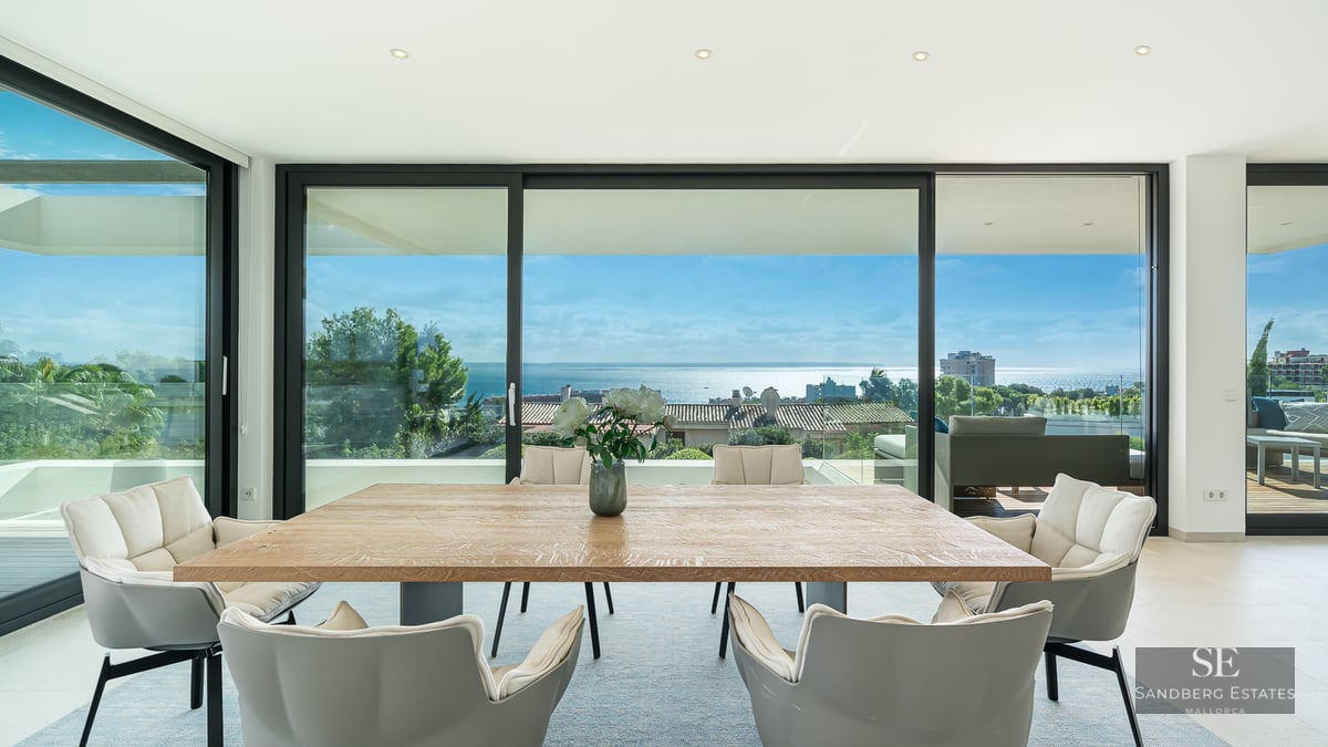 Bright dining room with a large wooden table and grey chairs facing floor-to-ceiling windows with a vast ocean view.