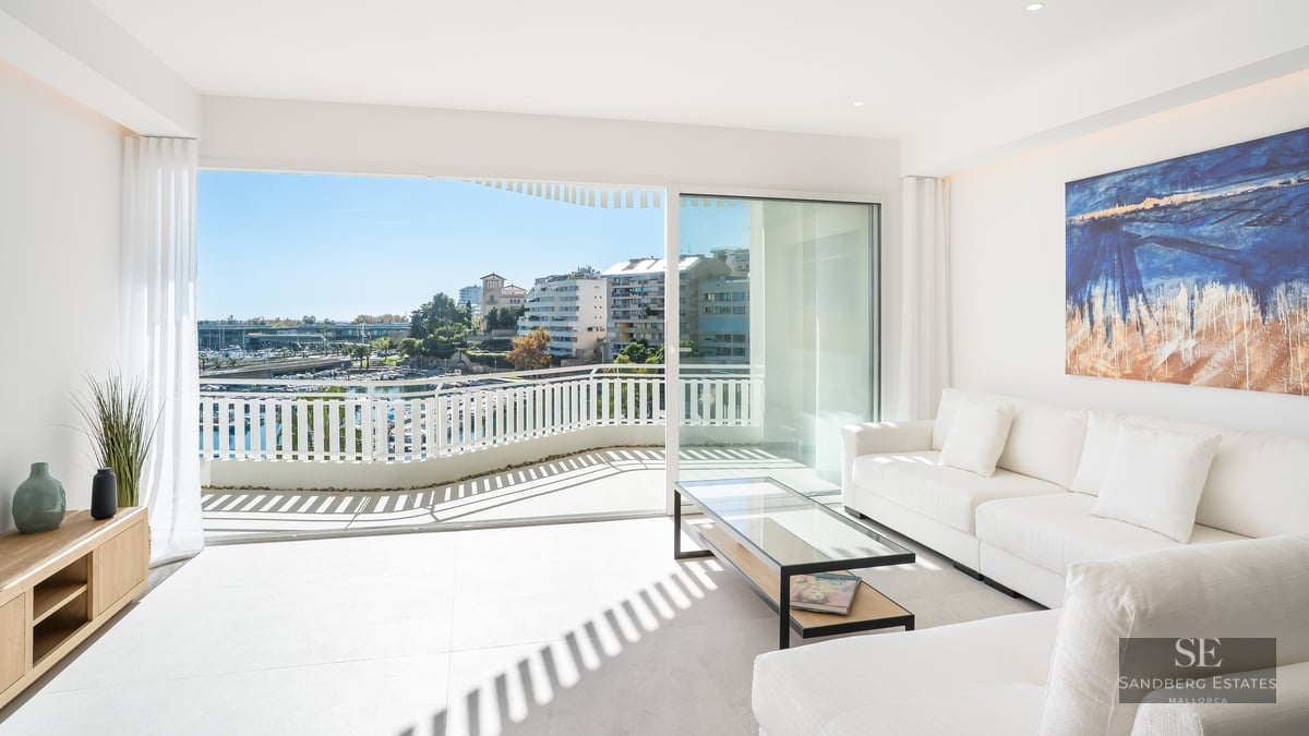 Bright living room with white sofas and glass doors opening to a balcony overlooking a marina.