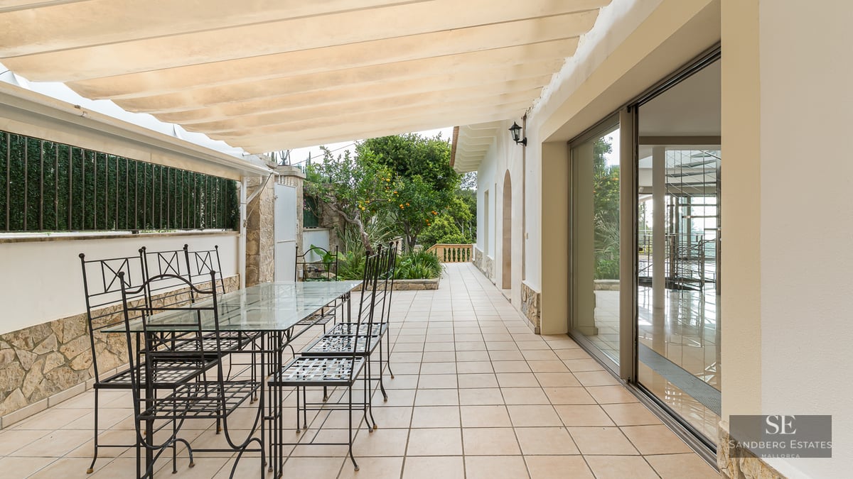 A shaded outdoor terrace with a glass dining table, black metal chairs, and a cream-colored retractable awning.