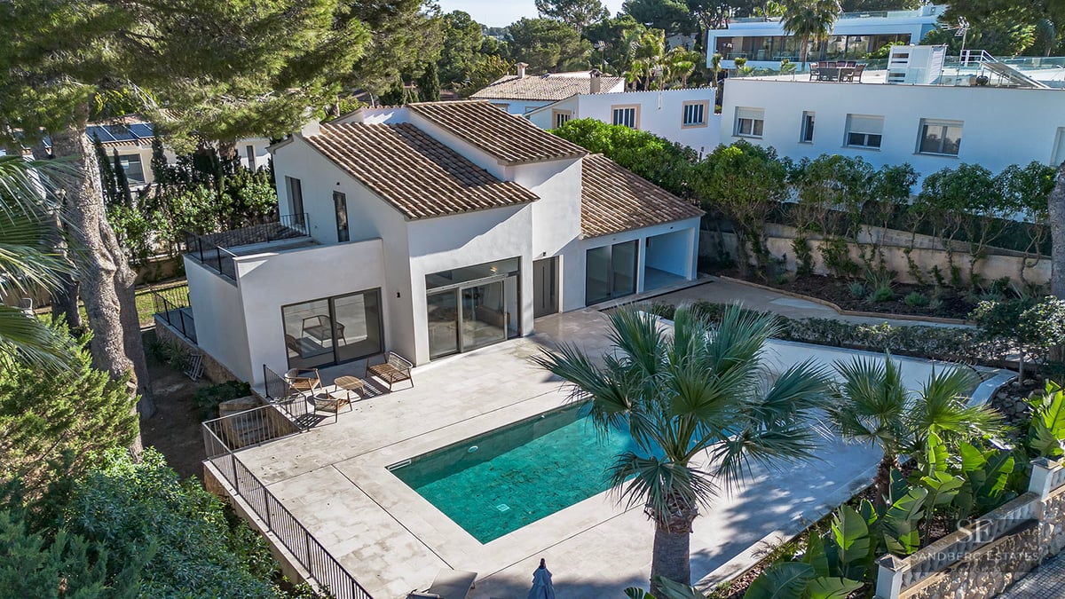 Aerial view of a white villa with a tiled roof, turquoise swimming pool, and large stone terrace surrounded by pine and palm trees.