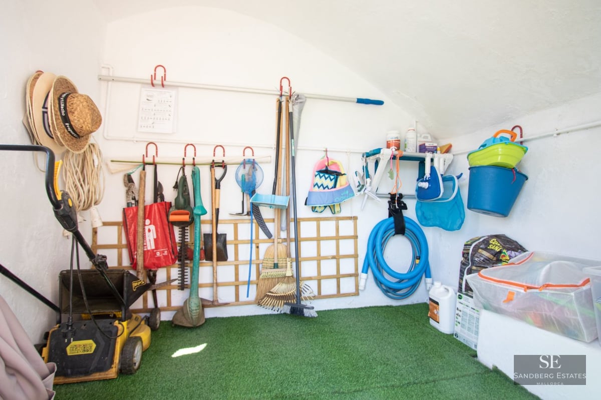 A white-walled storage room with artificial grass flooring containing a lawnmower, garden tools, and pool equipment.
