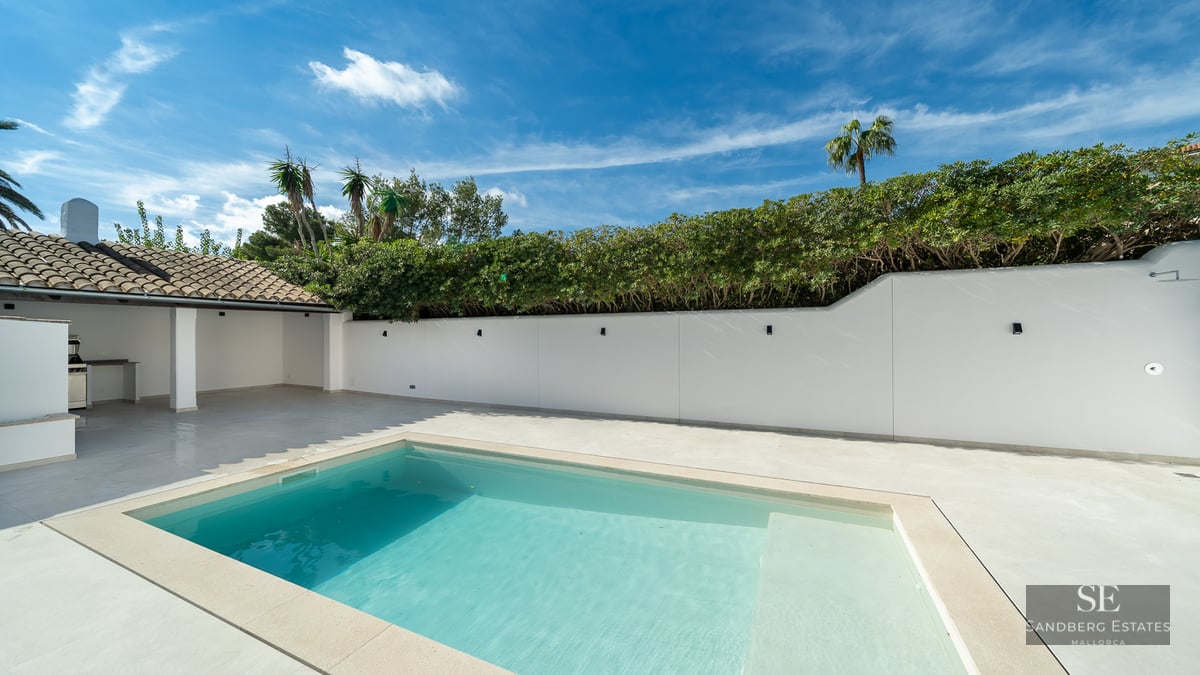 Turquoise swimming pool on a white stone terrace with a privacy wall, lush hedges, and an outdoor kitchen area.