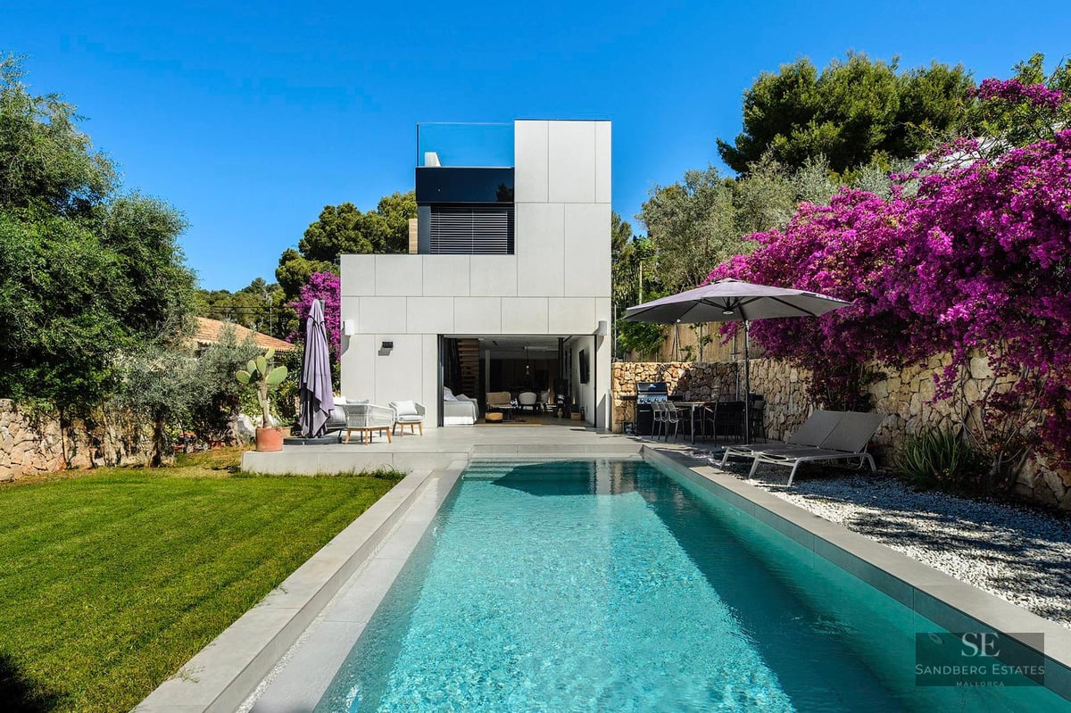 Modern white villa with a rectangular swimming pool, green lawn, and vibrant purple bougainvillea under a clear blue sky.