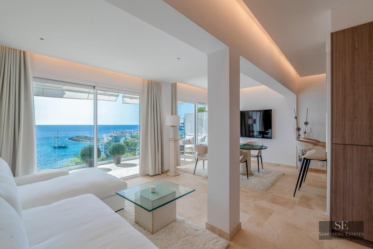 Bright modern living room with large glass doors opening to a Mediterranean sea view, featuring white sofas and stone floors.