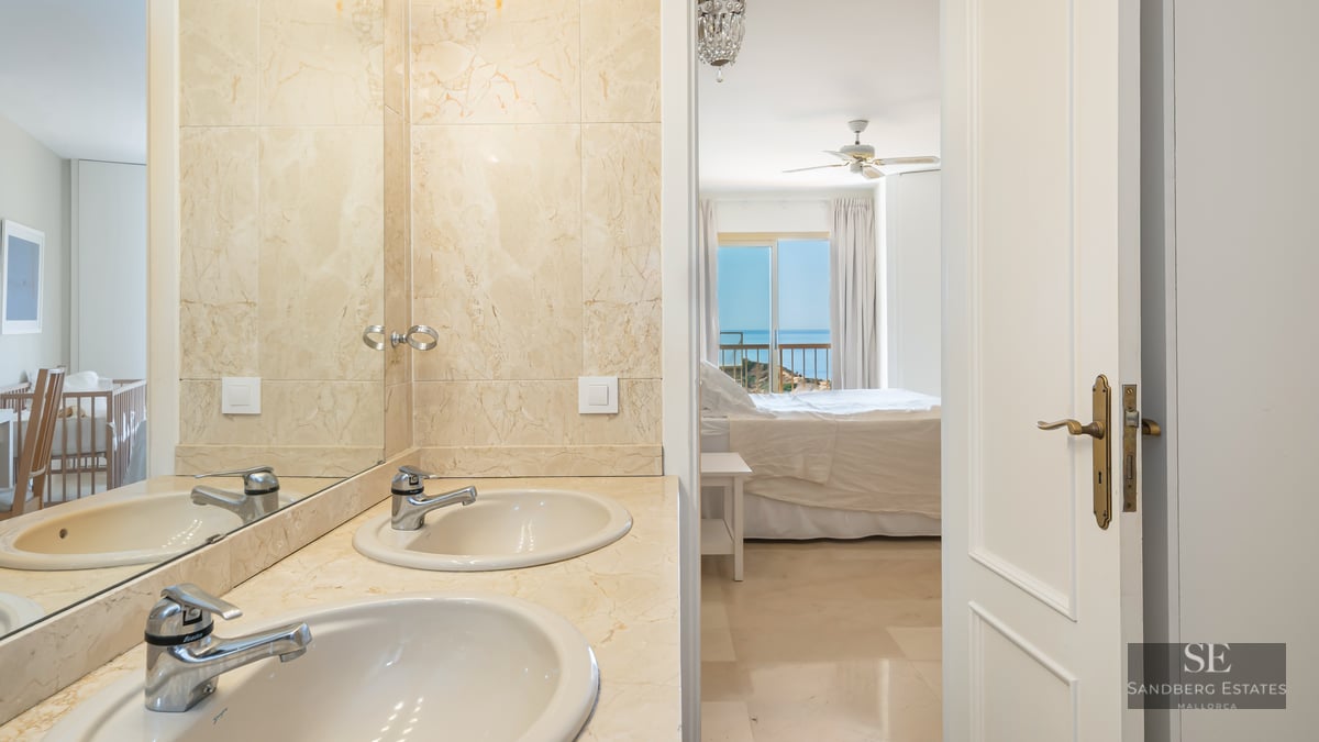 Master bathroom with beige marble double vanity looking into a bright bedroom with a sea view through an open door.