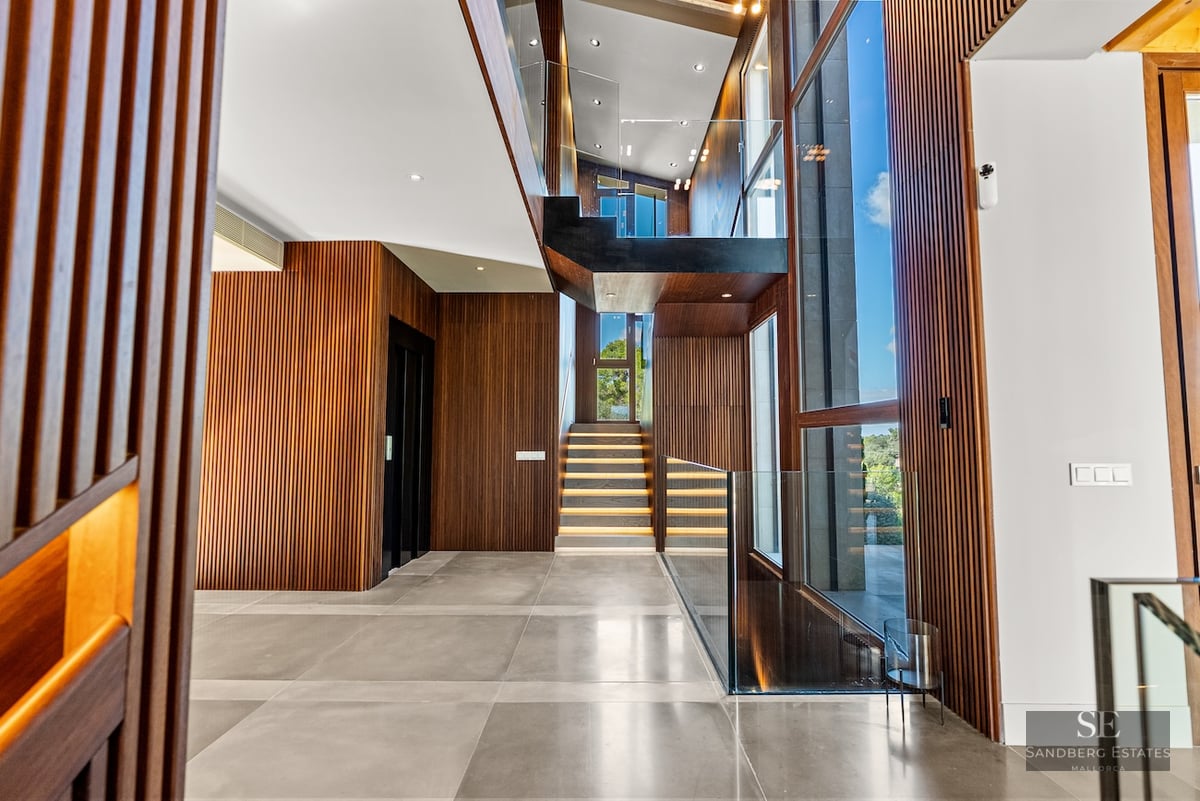 Modern foyer featuring slatted wood walls, a glass-railed staircase with LED lighting, and massive floor-to-ceiling windows.