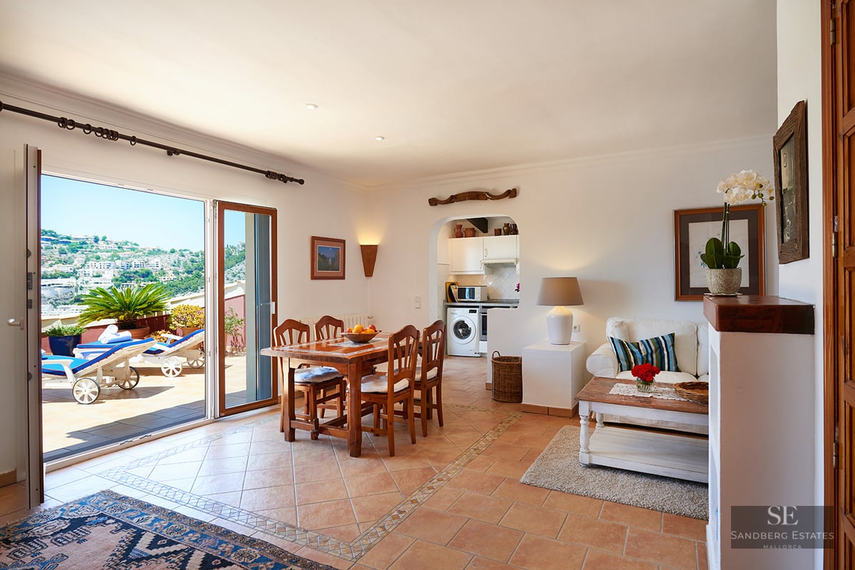 Bright open-plan living and dining room with terracotta floors opening onto a sunny terrace with hillside views.