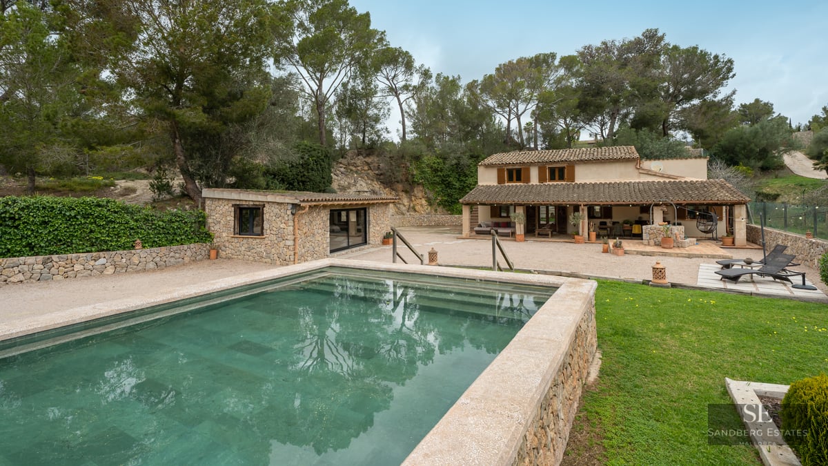 Outdoor swimming pool with stone borders in front of a rustic Mediterranean villa surrounded by pine trees.
