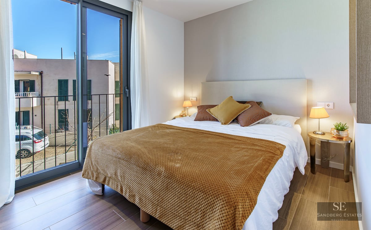 Bright bedroom featuring a double bed with an ochre yellow throw, a large glass sliding door, and a balcony view.