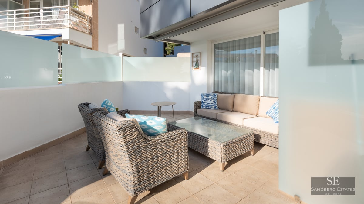 Terrasse moderne avec canapés en osier gris, coussins beige et bleus sur un patio ensoleillé avec parois en verre dépoli.