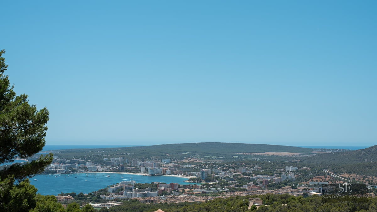 Panoramic elevated view of a blue Mediterranean bay, coastal town, and green hills under a clear blue sky.