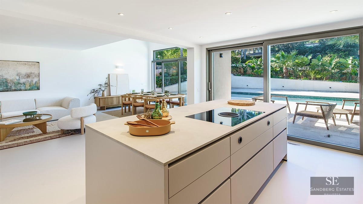 Bright, modern interior featuring a large kitchen island, dining area, sofas, and floor-to-ceiling windows leading to a pool.