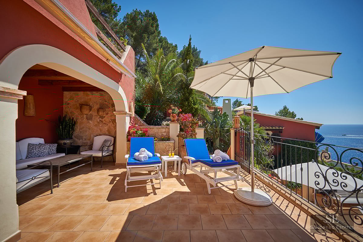 Sunny terrace with blue sun loungers, a white umbrella, and a wrought iron railing overlooking the Mediterranean Sea.